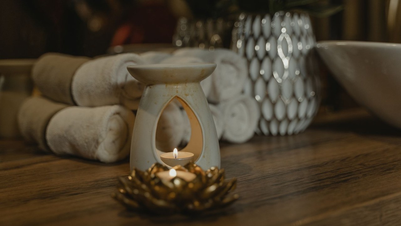 A lit candle in a ceramic warmer beside rolled white towels on a wood table